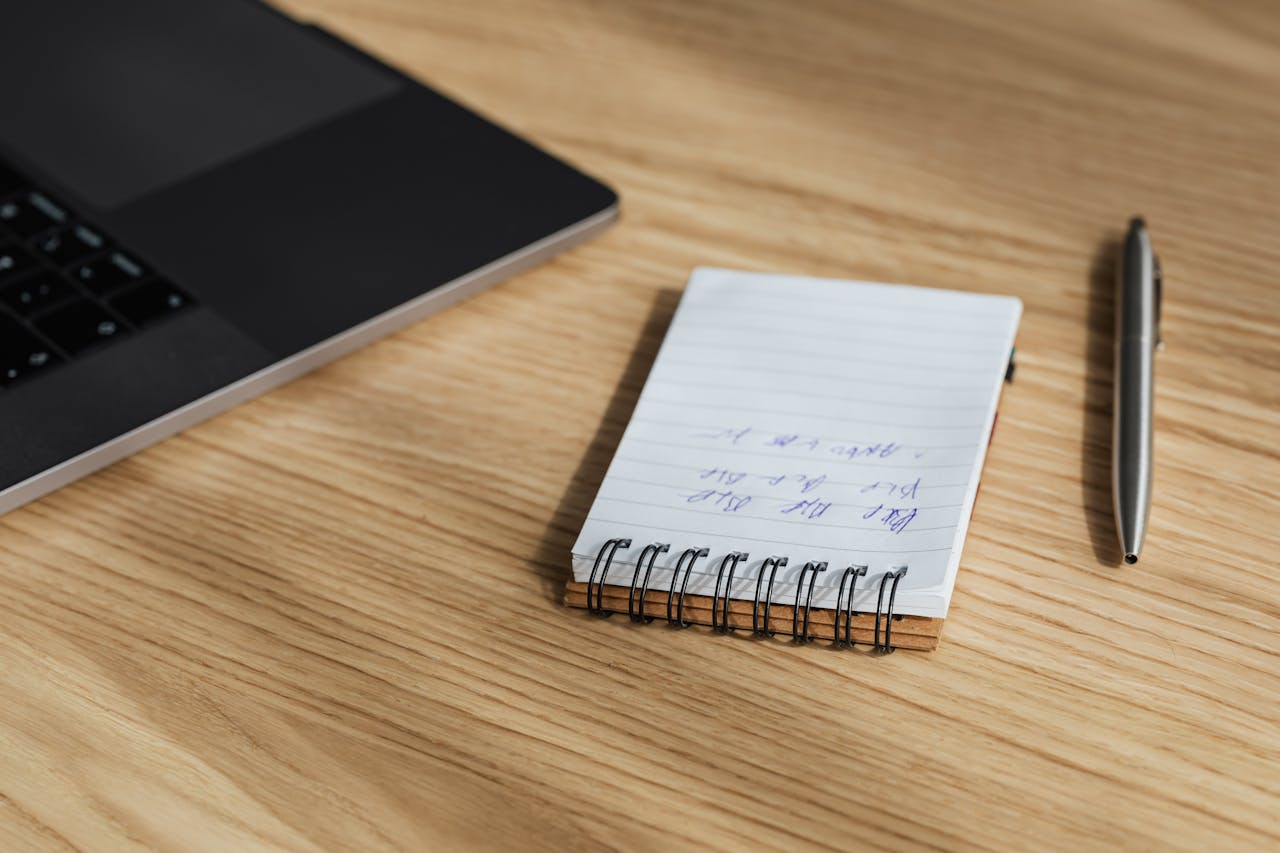 A wooden desk with a laptop, notepad with handwritten notes, and a pen, perfect for office work settings.